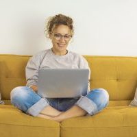 Happy young woman working on laptop sitting on sofa. Woman wokring from home. Young woman in eyeglasses smiling while working on laptop with crossed legs on sofa at home.